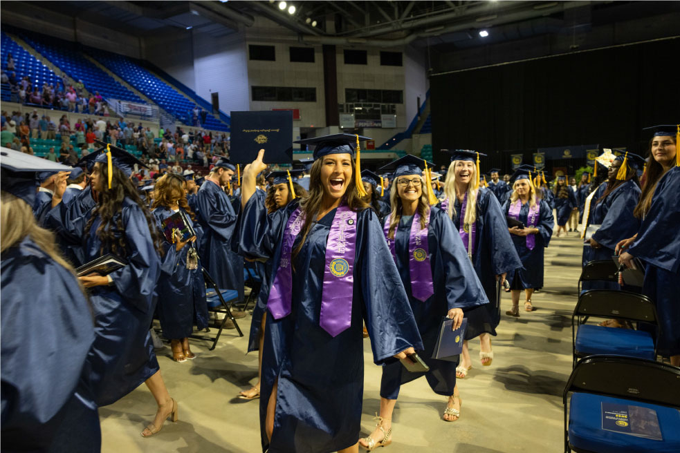 A student smiles with her college diploma at graduation.