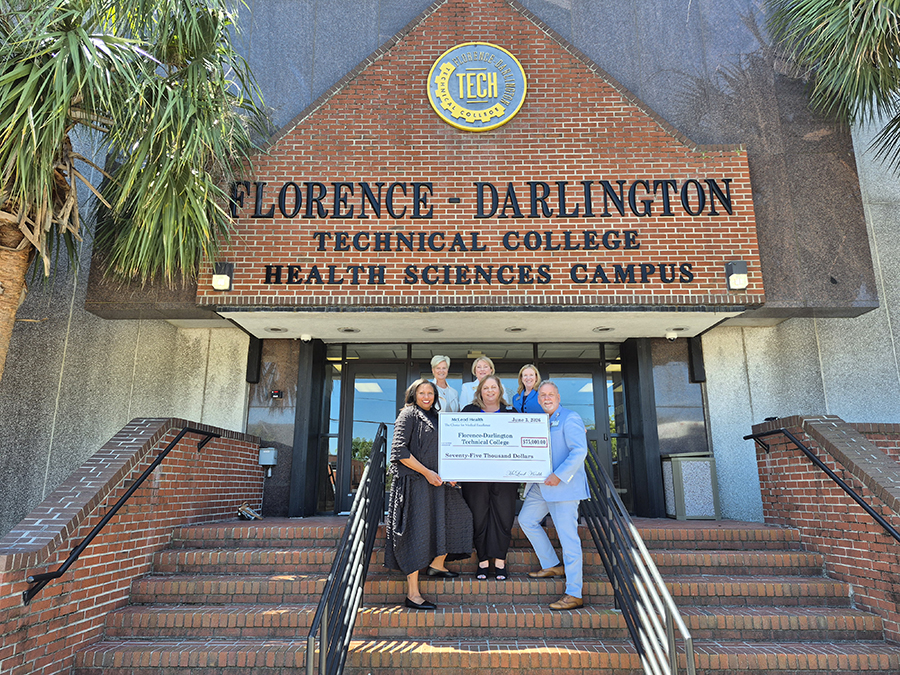 Six people stand on the steps of the FDTC Health Sciences Campus with a giant $75,000 donation from McLeod Health.