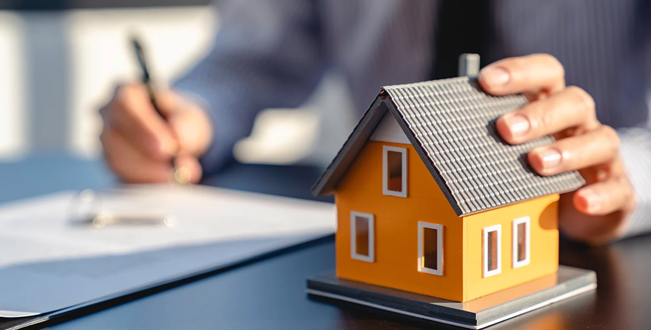 a real estate agent with one hand on a model house signs a contract with the other hand.